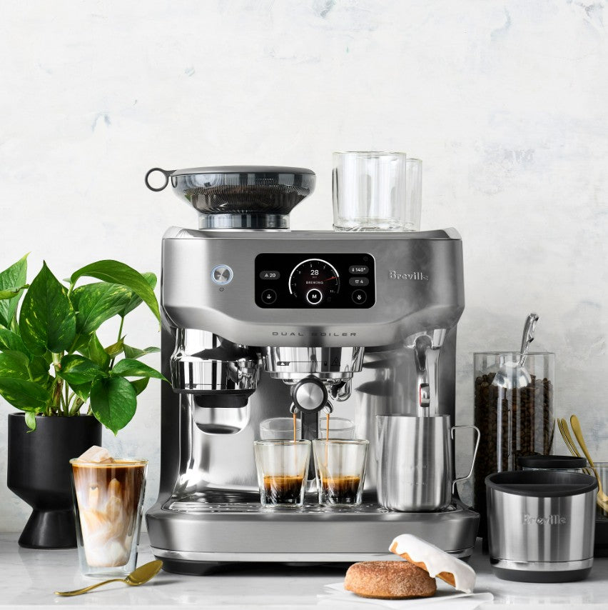 Breville coffee machine on a kitchen counter with glasses of coffee and a plant.