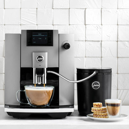 Jura coffee machine dispensing a cup of coffee with a black container and cookies on a white surface.