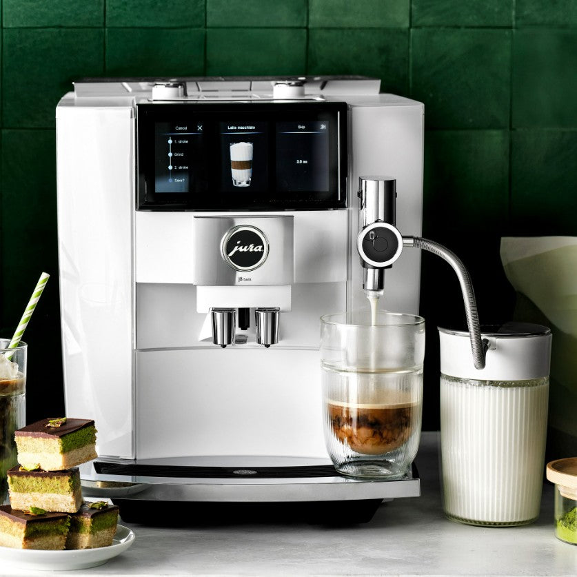 Silver coffee machine with a glass of coffee and cake on a green tiled wall background