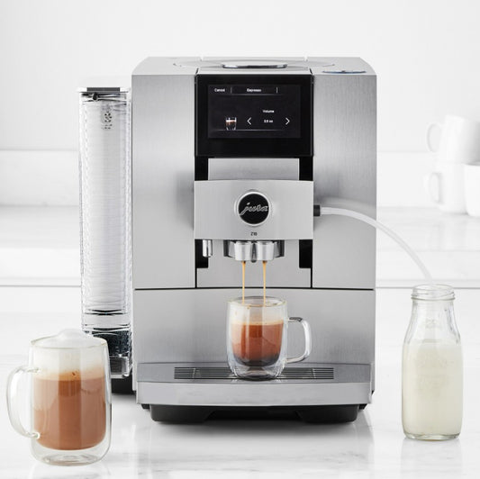 Silver coffee machine dispensing a cup of coffee on a white countertop with milk and another coffee cup nearby.