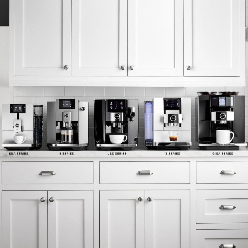 Row of coffee machines on a kitchen counter with white cabinets.