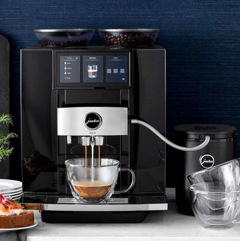 Black coffee machine with a glass cup of coffee on a countertop against a dark blue wall.