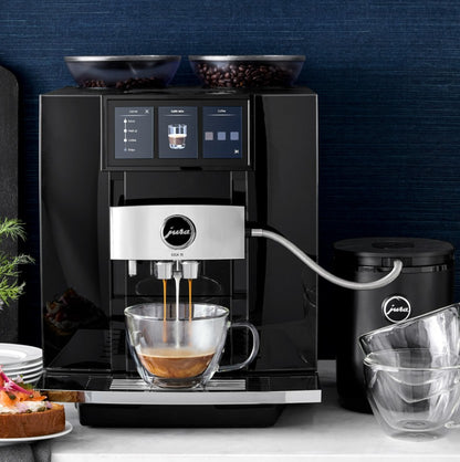 Black coffee machine with a glass cup of coffee on a countertop against a dark blue wall.