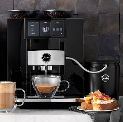 Coffee machine pouring coffee into a glass mug with a plate of food on a marble surface.